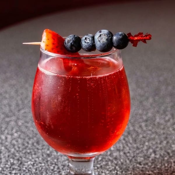Red cocktail in a wine glass, garnished with a strawberry and blueberries on a skewer.