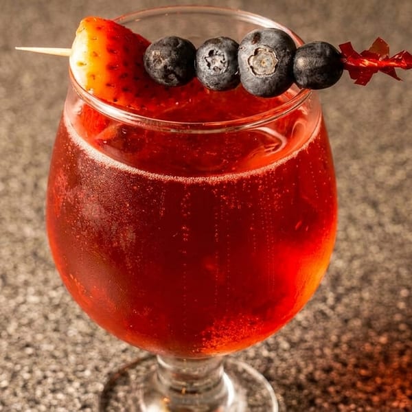Red sparkling drink in a glass, garnished with a strawberry and blueberries on a skewer.