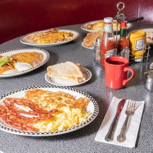 Diner table laden with multiple breakfast plates of eggs, bacon, hash browns, and toast.