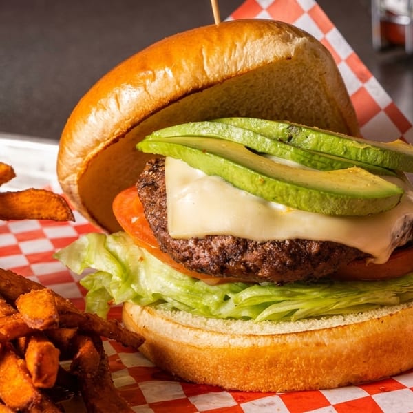 A juicy avocado cheeseburger with lettuce and tomato, served with sweet potato fries on a metal tray.