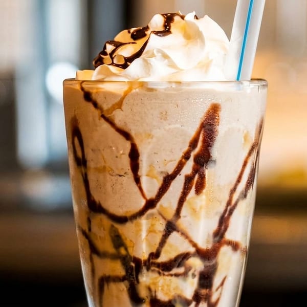 A tall glass of milkshake with whipped cream, chocolate drizzle, and a blue-striped straw.