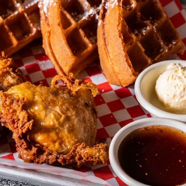 Crispy fried chicken and waffles with powdered sugar, butter, and syrup on a checkered paper-lined tray.